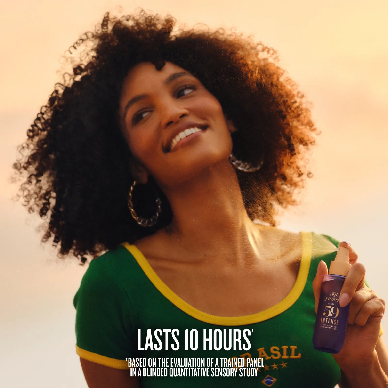 A woman with curly hair wearing a green and yellow top holds a purple bottle of Dr. Brandis 30 Intensity SPF 50 sunscreen against a warm sunset sky.