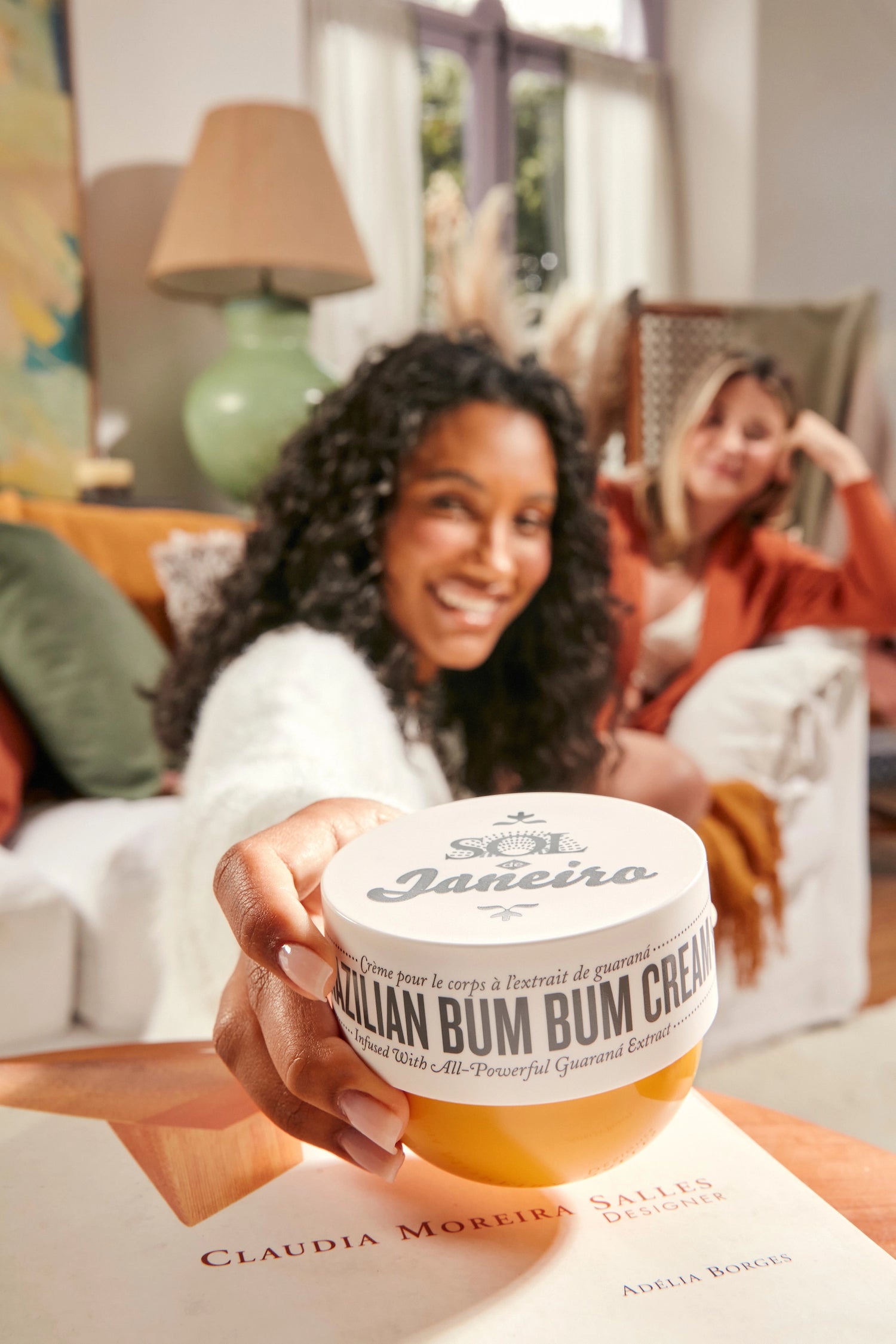 two women sitting on a couch. One one is reaching for a jar of Brazilian Bum Bum Cream