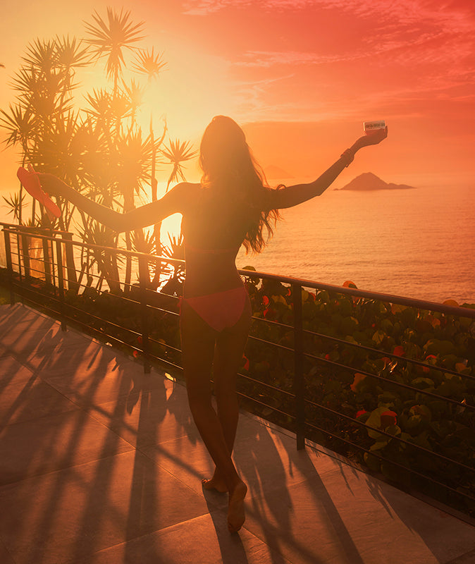 Woman with her arms wide holding Bom Dia Bright Body Cream and facing the sun