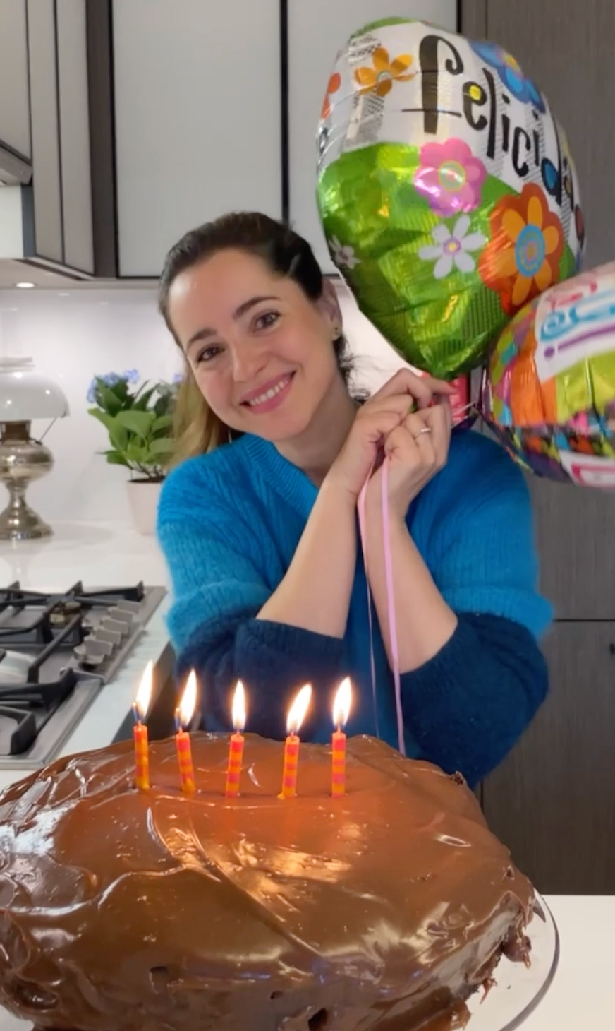 woman in kitchen holding birthday balloons and smiling with a birthday cake in front of her with 5 candles