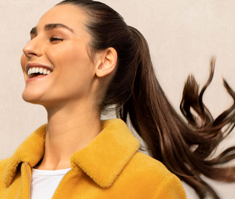 Woman with flowing straight hair smiling and wearing a yellow coat on a beige background