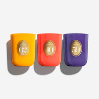 Three Noël Enroçé candle jars in bright yellow, coral red, and deep purple, arranged side by side on a light gray background with soft shadows.