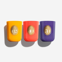 Three Noël Enroçé candle jars in bright yellow, coral red, and deep purple, arranged side by side on a light gray background with soft shadows.