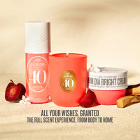 Three Sol Jenévro products arranged together on a warm beige surface: a pink body mist bottle, a coral candle, and a pink room spray jar with white lids, styled with red fruits, white flowers, and wooden sticks. Text at the bottom reads, 'ALL YOUR WISHES, GRANTED THE FULL SCENT EXPERIENCE, FROM BODY TO HOME.