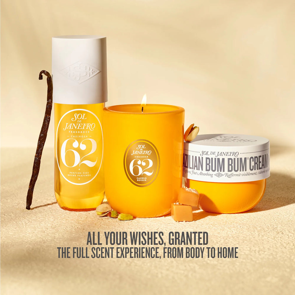 Three Sol Janeiro 62 scented products arranged on a sandy beige surface: a tall clear bottle with a white cap, a lit orange-yellow candle, and an orange-yellow cream jar labeled 'Italian Bum Bum Cream,' with a vanilla bean and small nuts nearby. Text at the bottom reads, 'ALL YOUR WISHES, GRANTED THE FULL SCENT EXPERIENCE, FROM BODY TO HOME.