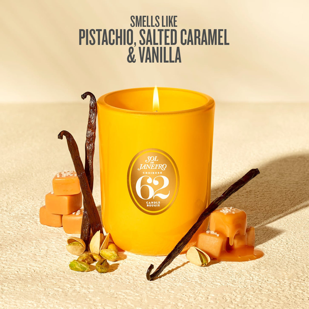 A yellow candle with a lit wick sits on a textured beige surface, surrounded by vanilla beans, pistachios, caramel cubes, and caramel drizzle. Text at the top reads 'SMELLS LIKE PISTACHIO, SALTED CARAMEL & VANILLA.