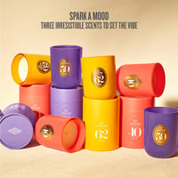 A group of colorful cylindrical candle containers in purple, yellow, and coral red, arranged on a beige surface with the text 'SPARK A MOOD THREE IRRESISTIBLE SCENTS TO SET THE VIBE' above them. Each container has a round gold label with a number and the brand name 'SPA JANSIRO'.
