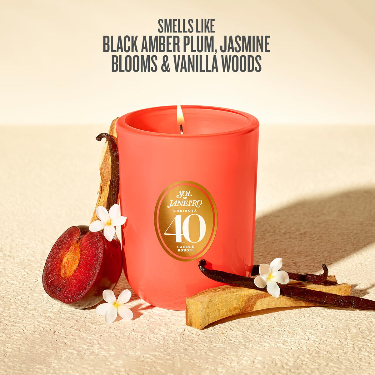 A red-orange scented candle in a matte jar with a gold circular label reading 'Sol Janeiro Brazil 40 Anos'. The candle is styled on a textured beige surface with a sliced plum, vanilla beans, and small white flowers, with text above describing the scent notes.