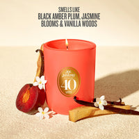 A red-orange scented candle in a matte jar with a gold circular label reading 'Sol Janeiro Brazil 40 Anos'. The candle is styled on a textured beige surface with a sliced plum, vanilla beans, and small white flowers, with text above describing the scent notes.