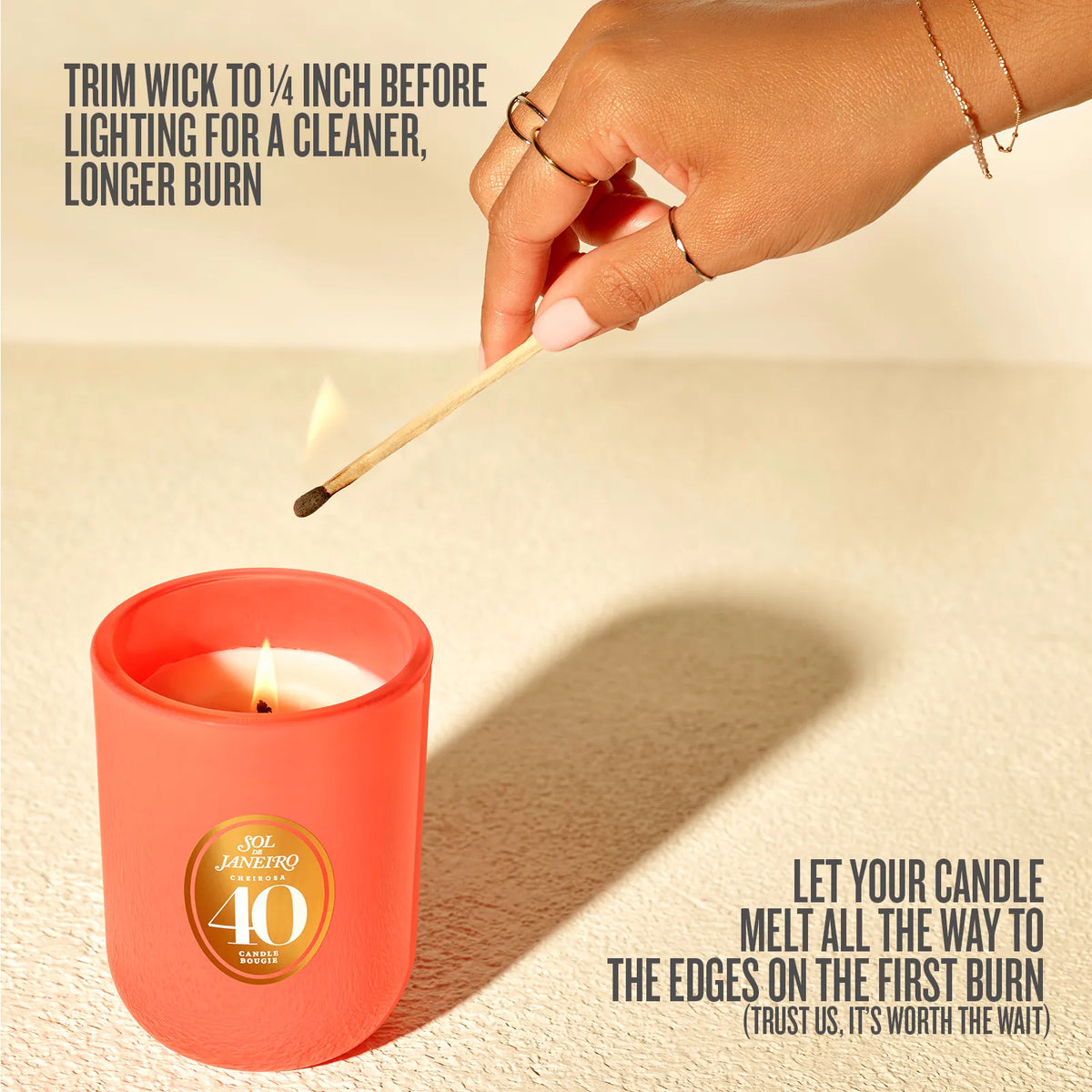 A coral pink glass candle jar with a gold circular '40th Anniversary' logo, shown on a textured beige surface with a hand lighting a match above it. Text overlays explain care instructions for the candle burn.