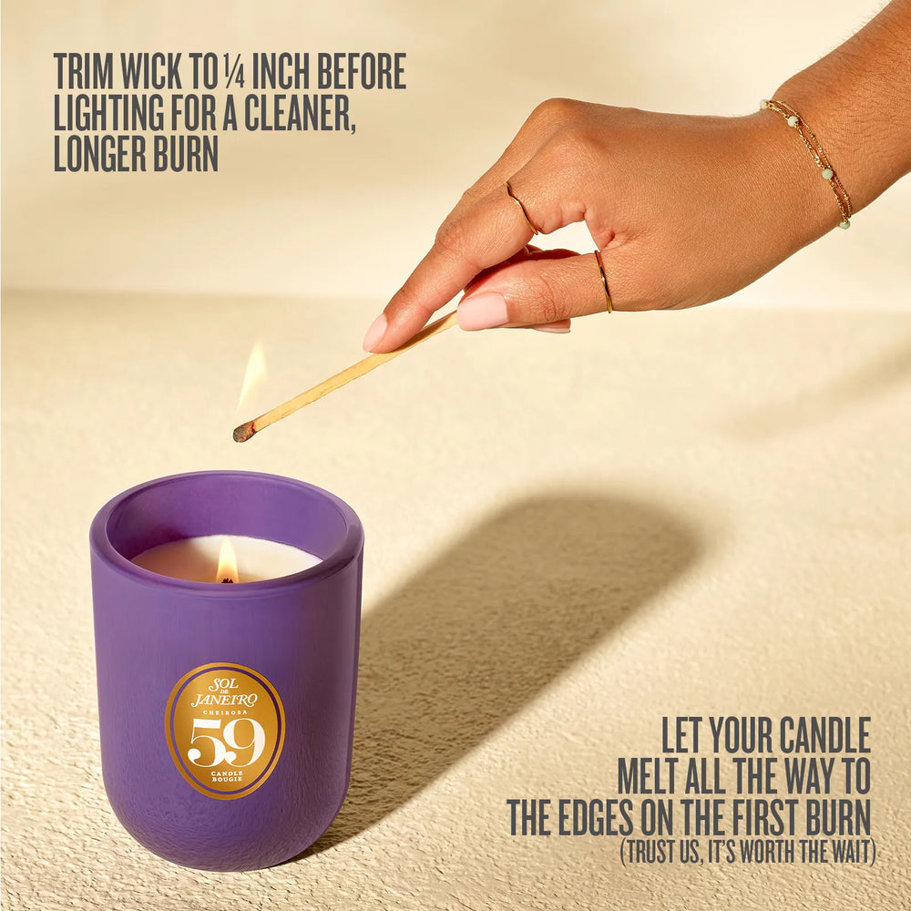 A lit purple candle in a matte container with a gold circular label reading 'You Are Loved 59', shown on a beige textured surface with a hand lighting it with a match. Text overlays describe candle care instructions.