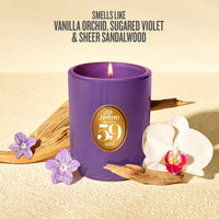 A purple soy candle with a gold circular label reading 'Sol de Janeiro 59' is styled on a beige textured surface with driftwood, purple flowers, and a white orchid. Text at the top reads, 'SMELLS LIKE VANILLA ORCHID, SUGARED VIOLET & SHEER SANDALWOOD'.