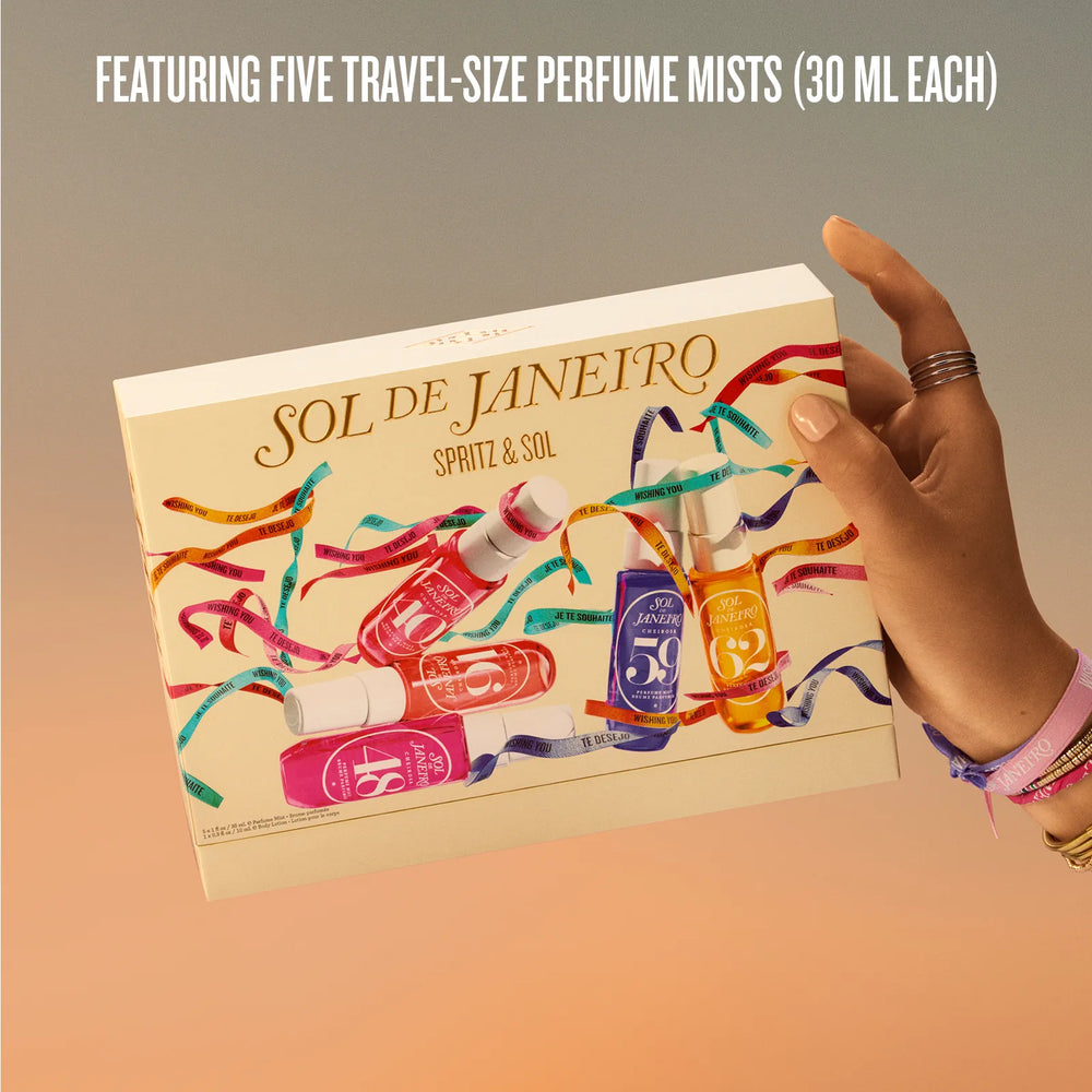 A hand holds a beige box of SOL DE JANEIRO Spritz & Sol travel-size perfume mists against a warm gradient background. The box features colorful illustrations of miniature perfume bottles wrapped in multicolored streamers, with text at the top reading 'FEATURING FIVE TRAVEL-SIZE PERFUME MISTS (30 ML EACH)'.