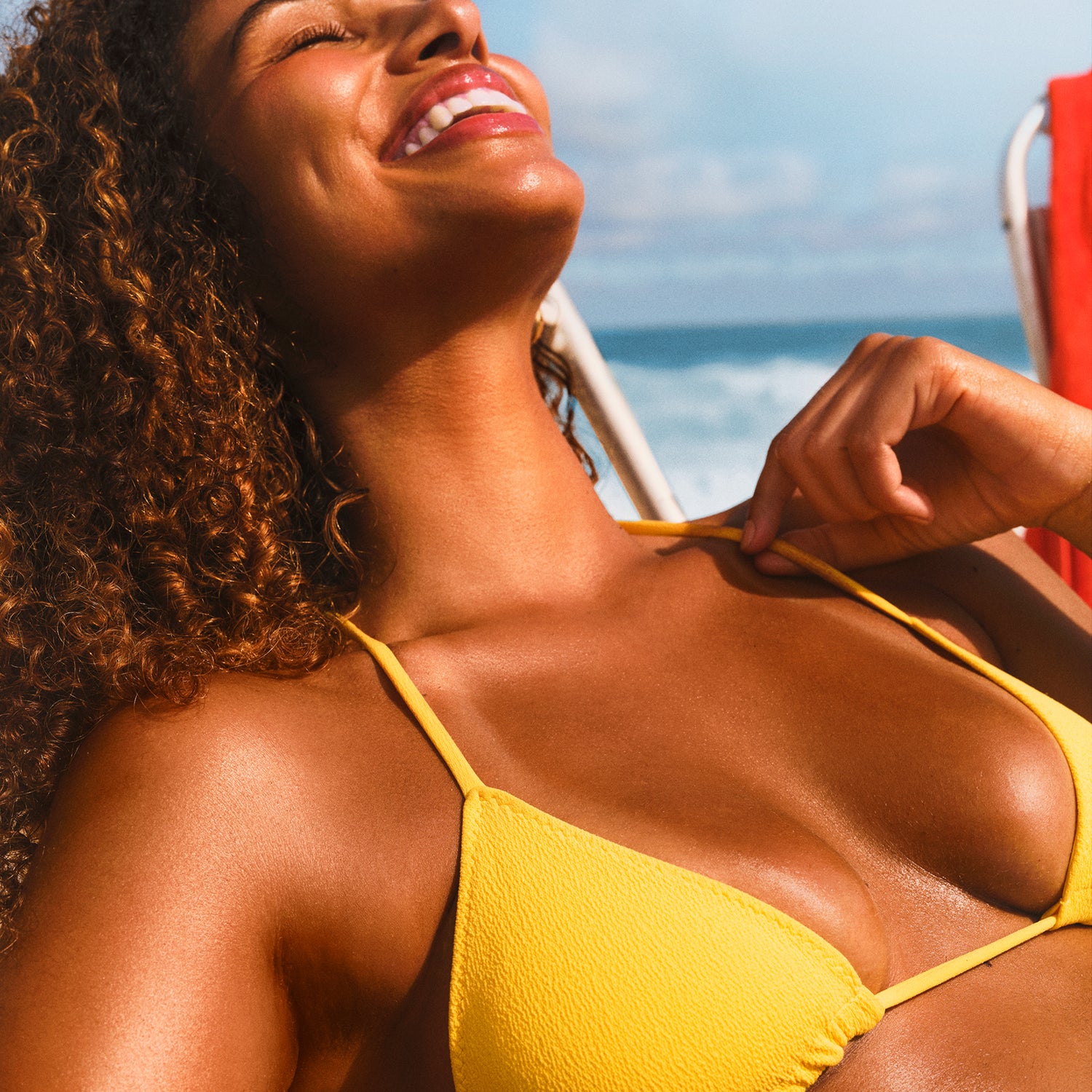 Curly haired model smiling on beach in sun with a yellow bikini.