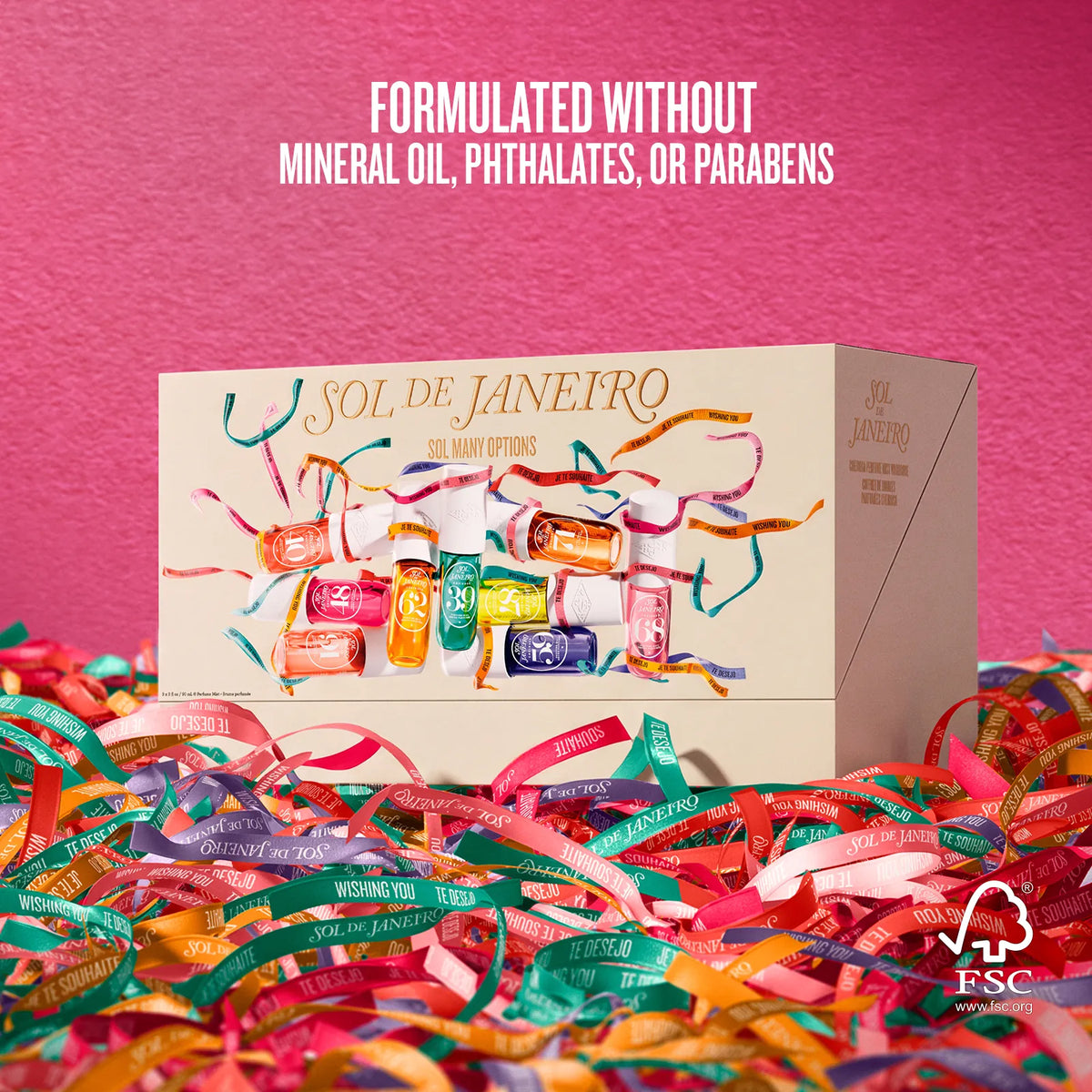 A boxed cosmetic set labeled SOL DE JANEIRO sits on a bed of colorful ribbon against a bright pink background. Text at the top reads 'FORMULATED WITHOUT MINERAL OIL, PHTHALATES, OR PARABENS.