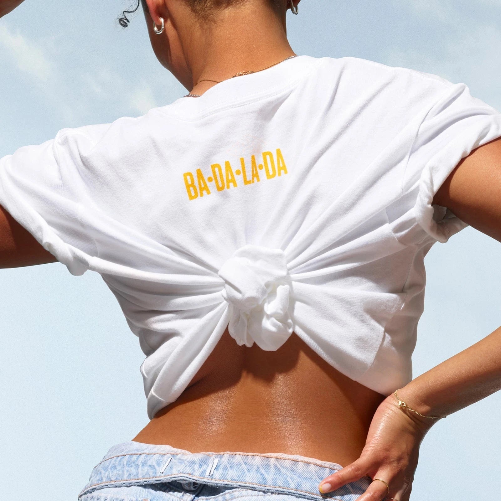 sol de janeiro badalada t-shirt, on the back of t-shirt it reads "ba-da-la-da" in sol de janeiro yellow. T shirt is white
