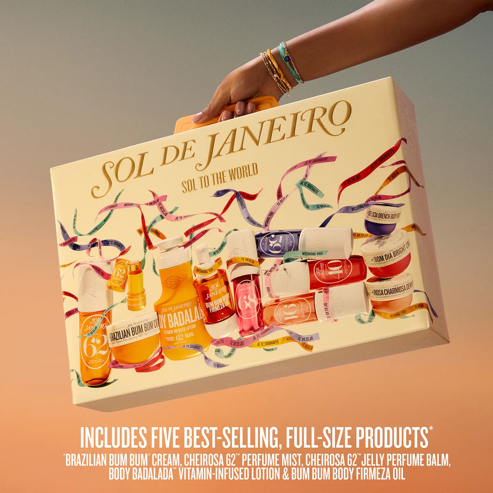 A hand holds a large Sol de Janeiro gift box against a warm beige-to-orange gradient background. The cream-colored box features the brand name in gold script and colorful illustrations of the included products, including Brazilian Bum Bum Cream, Cheirusa 62, Jelly Perfume Balm, Body Badalada, and Vitamin-Infused Lotion and Body Fierce Oil.