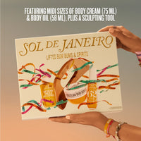 A person holds a boxed body care set labeled SOL DE JANEIRO against a soft beige-to-gray gradient background. The box features illustrated products including a body cream jar, body oil bottle, and a sculpting tool, with colorful ribbon graphics and the text 'FEATURING MIDI SIZES OF BODY CREAM (75 ML) & BODY OIL (58 ML), PLUS A SCULPTING TOOL.