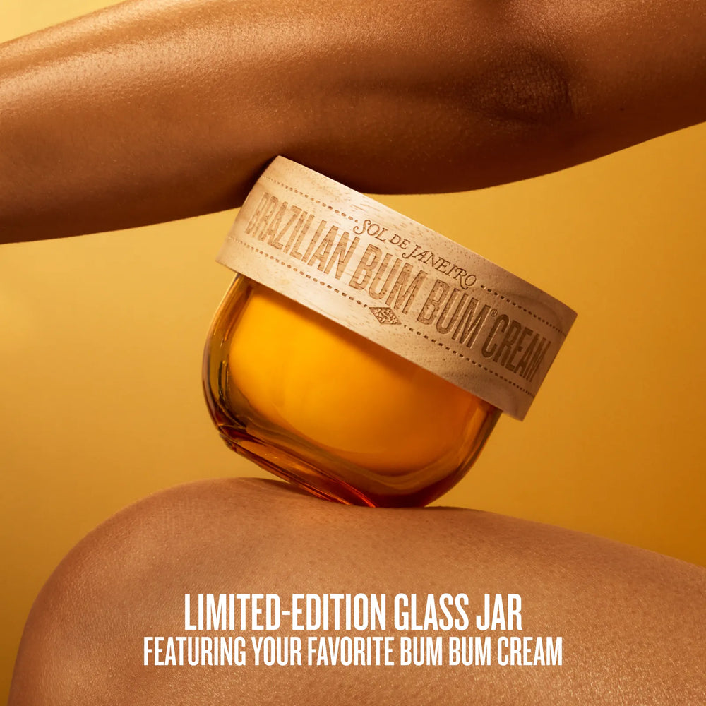 A limited-edition glass jar of Sol de Janeiro Brazilian Bum Bum Cream resting on a model's lap, with the model's arms framing the product against a warm golden-yellow background.