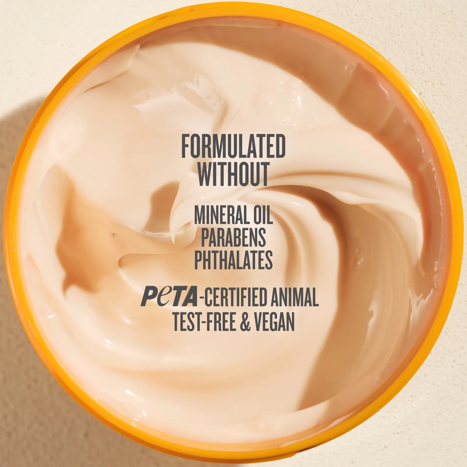 Top-down view of a beige cosmetic cream in a round tub with a yellow-orange rim, featuring text that reads 'FORMULATED WITHOUT MINERAL OIL PARABENS PHTHALATES PETA-CERTIFIED ANIMAL TEST-FREE & VEGAN'.