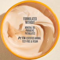 Top-down view of a beige cosmetic cream in a round tub with a yellow-orange rim, featuring text that reads 'FORMULATED WITHOUT MINERAL OIL PARABENS PHTHALATES PETA-CERTIFIED ANIMAL TEST-FREE & VEGAN'.