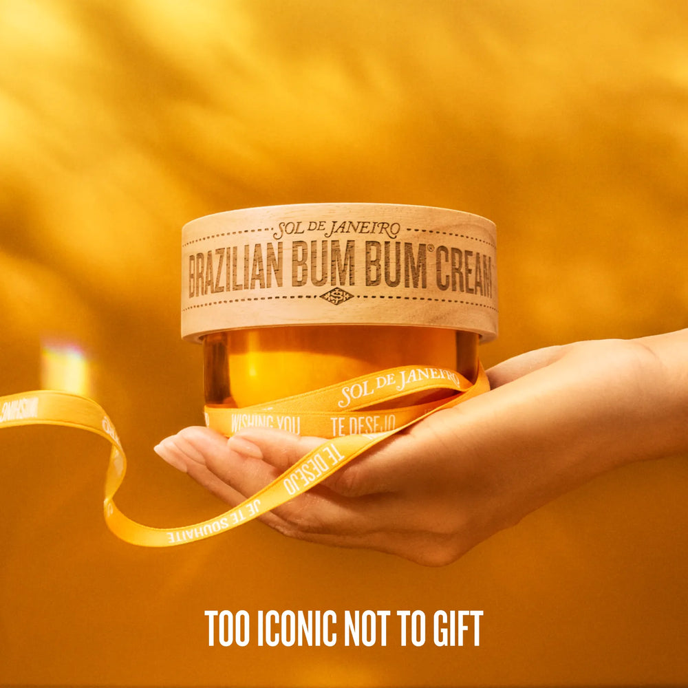 A hand holding a jar of Sol de Janeiro Brazilian Bum Bum Cream against a warm golden-yellow background, wrapped in a matching yellow ribbon with the brand name printed repeatedly. White text at the bottom reads 'TOO ICONIC NOT TO GIFT'.