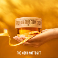 A hand holding a jar of Sol de Janeiro Brazilian Bum Bum Cream against a warm golden-yellow background, wrapped in a matching yellow ribbon with the brand name printed repeatedly. White text at the bottom reads 'TOO ICONIC NOT TO GIFT'.
