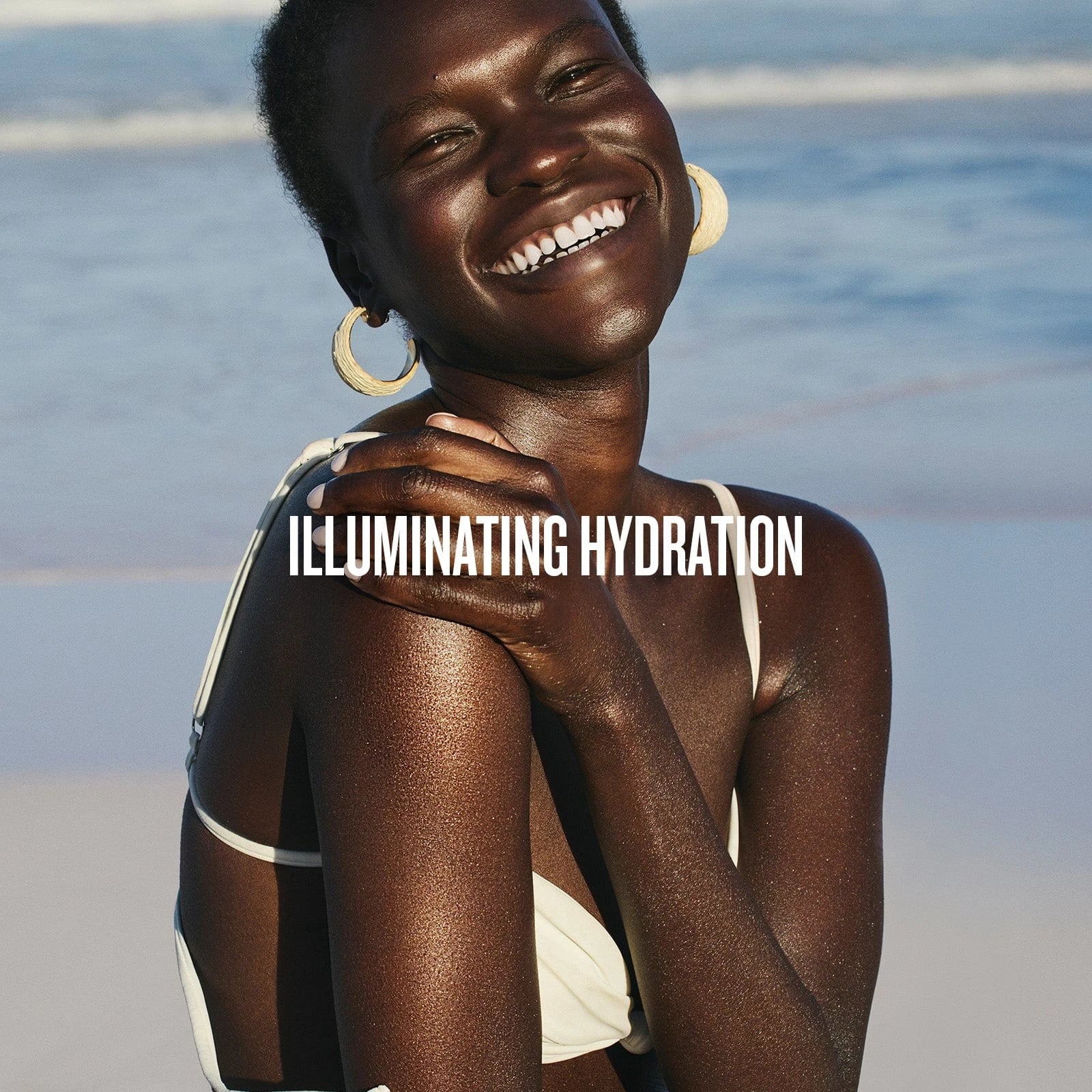 text on image reads illuminating hydration. This image is of a model on the beach with the ocean in backgound smiling and looking glowy wearing sol de janeiro glowmotions glow body oil