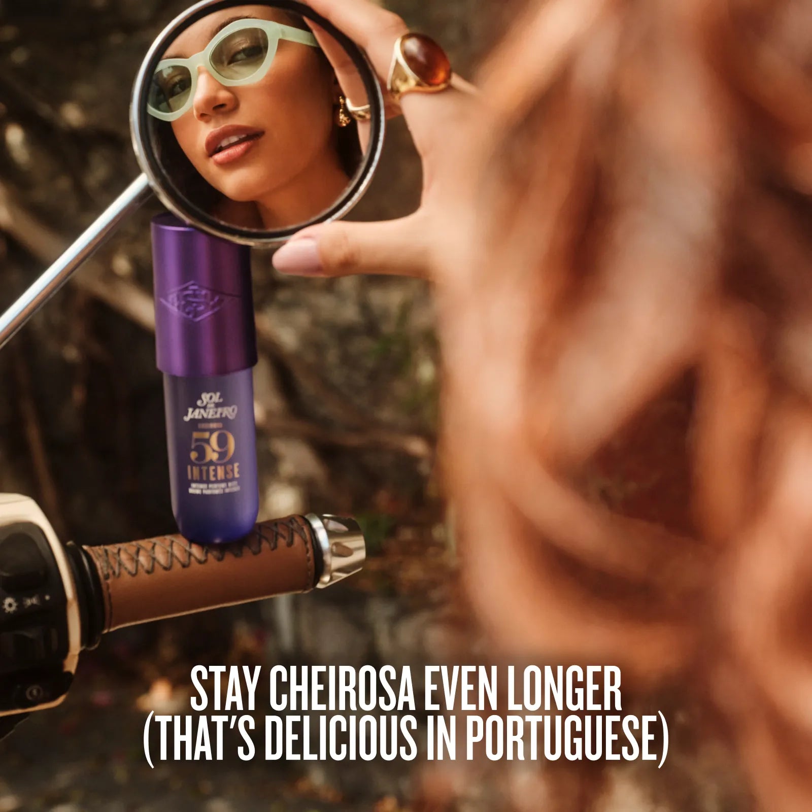A close-up lifestyle shot of a person holding a round mirror to inspect a purple tube of Cheirosome 59 Intense foundation. The background is heavily blurred, showing warm skin tones and brown hair, with a motorcycle handlebar visible on the left.