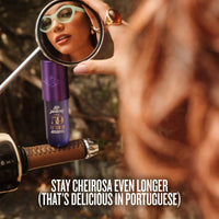 A close-up lifestyle shot of a person holding a round mirror to inspect a purple tube of Cheirosome 59 Intense foundation. The background is heavily blurred, showing warm skin tones and brown hair, with a motorcycle handlebar visible on the left.