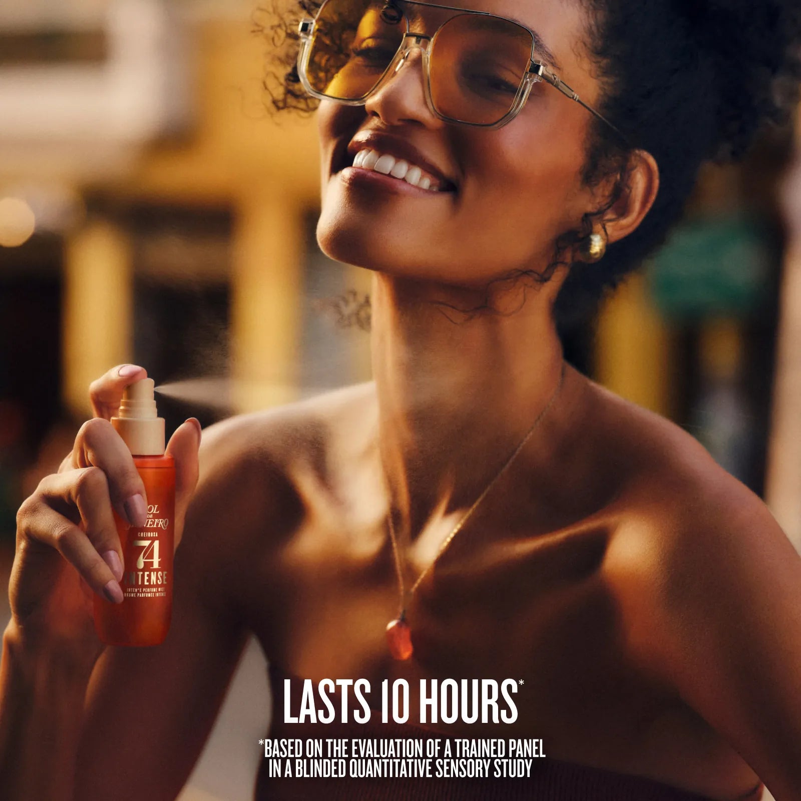 A woman wearing large tinted sunglasses and a gold necklace sprays a bottle of orange sunscreen on her neck in an outdoor setting. Text at the bottom reads &