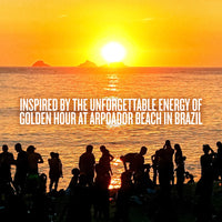 A golden sunset over the ocean with silhouetted people standing along the shoreline, and large overlaid text reading: 'INSPIRED BY THE UNFORGETTABLE ENERGY OF GOLDEN HOUR AT ARPOADOR BEACH IN BRAZIL.'.