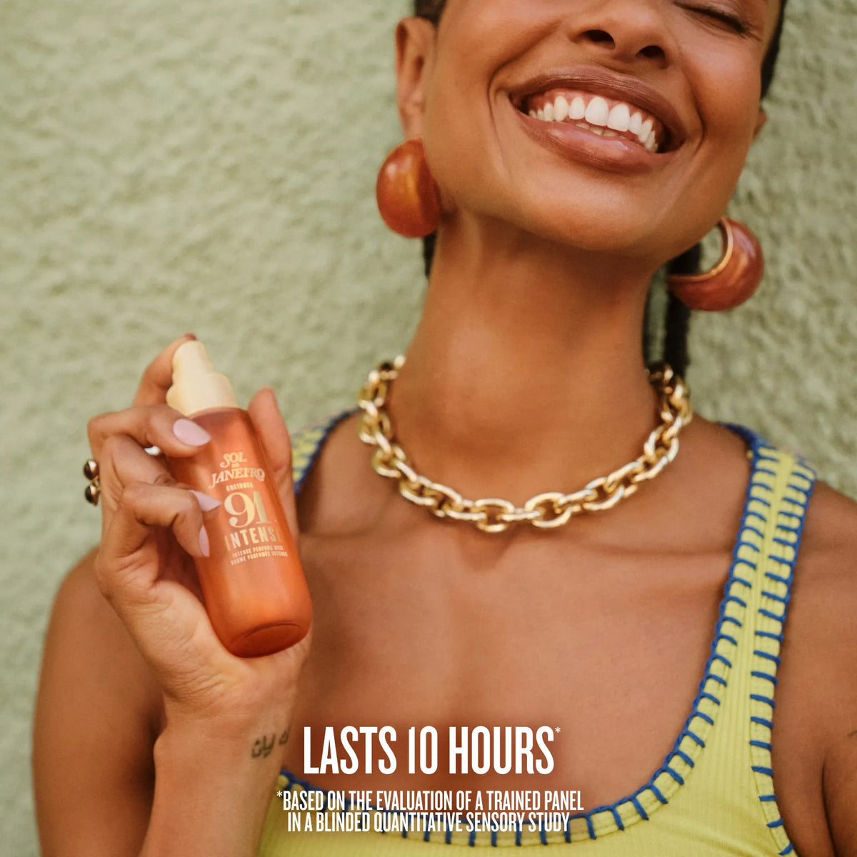 A smiling woman holds up a small orange-tinted bottle of Solde Janeiro Cheirosa 91 Intense Perfume Mist against a textured light green wall. The image features promotional text at the bottom stating 'LASTS 10 HOURS'. *BASED ON THE EVALUATION OF A TRAINED PANEL IN A BLINDED QUANTITATIVE SENSORY STUDY