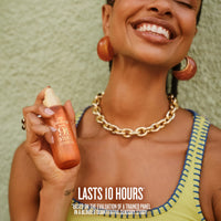 A smiling woman holds up a small orange-tinted bottle of Solde Janeiro Cheirosa 91 Intense Perfume Mist against a textured light green wall. The image features promotional text at the bottom stating 'LASTS 10 HOURS'. *BASED ON THE EVALUATION OF A TRAINED PANEL IN A BLINDED QUANTITATIVE SENSORY STUDY