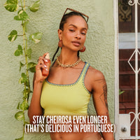A woman wearing a lime green tank top and gold jewelry holds a bottle of Sol de Janeiro chairosa 91 perfume mist against a textured green wall with a hanging plant. Text at the bottom reads 'STAY CHEIROSA EVEN LONGER (THAT'S DELICIOUS IN PORTUGUESE)'.