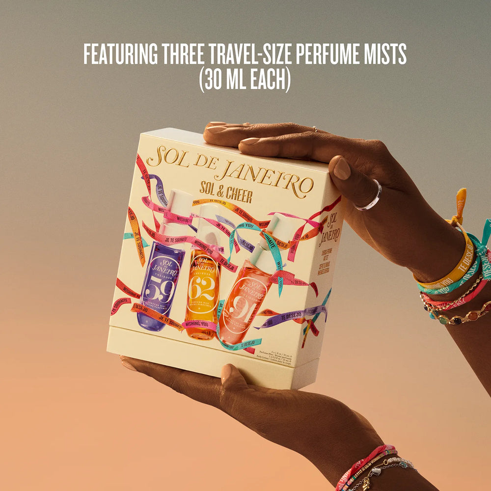 A person holds a Sol de Janeiro travel-size perfume mist gift box featuring three 30 ml mists. The cream-colored box is decorated with colorful confetti and illustrations of three perfume bottles (purple, orange, and pink).
