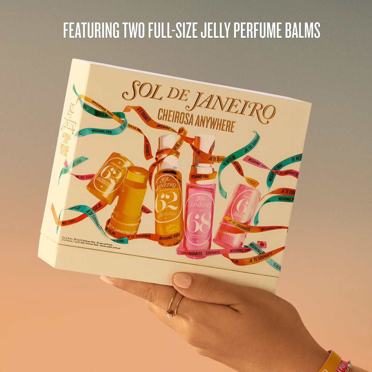 A hand holds a cream-colored box of Sol de Janeiro Cheirosa Anywhere perfume balms against a warm gradient background. The box features colorful illustrations of the balm bottles and decorative ribbons, with text at the top reading 'FEATURING TWO FULL-SIZE JELLY PERFUME BALMS'.